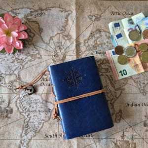 May include: A blue leather journal with a compass design and a brown elastic band sits on a vintage world map. A pink flower, Euro banknotes, and coins are also on the map, suggesting travel and exploration.