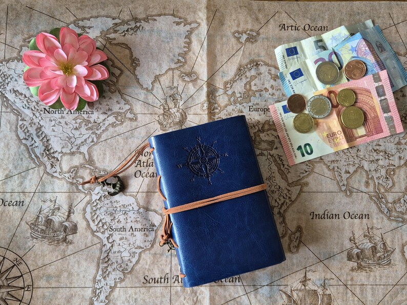 May include: A blue leather journal with a compass design and a brown elastic band sits on a vintage world map. A pink flower, Euro banknotes, and coins are also on the map, suggesting travel and exploration.