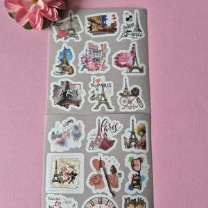 May include: Two sheets of Paris-themed stickers featuring the Eiffel Tower, fashion illustrations, and the word "Paris" in various fonts. The stickers are in shades of pink, white, and black. A pink flower is in the upper left corner.