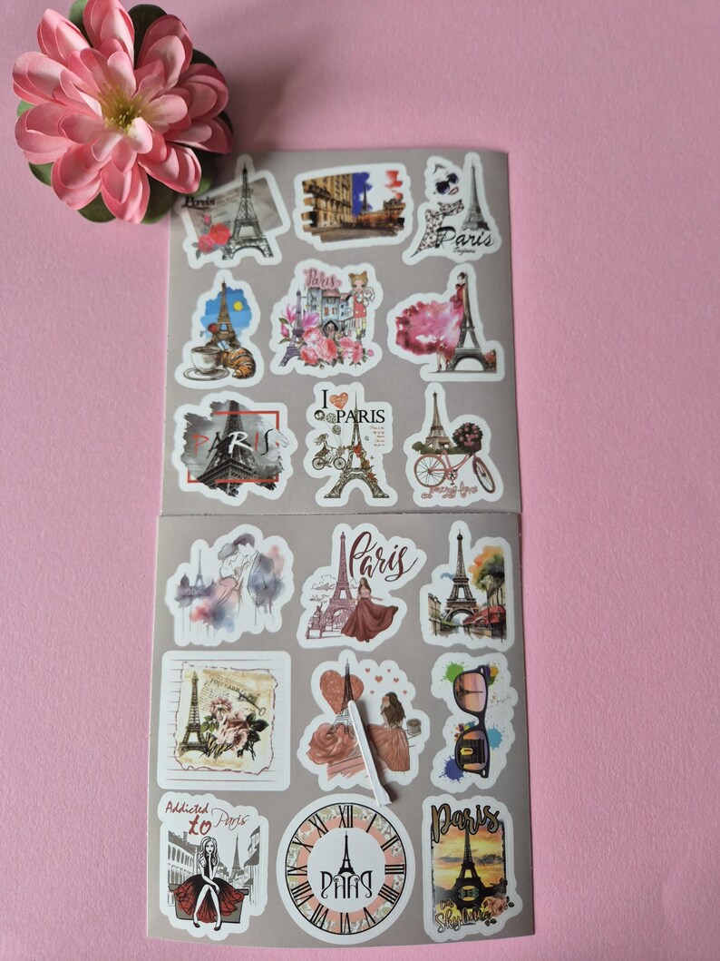 May include: Two sheets of Paris-themed stickers featuring the Eiffel Tower, fashion illustrations, and the word "Paris" in various fonts. The stickers are in shades of pink, white, and black. A pink flower is in the upper left corner.