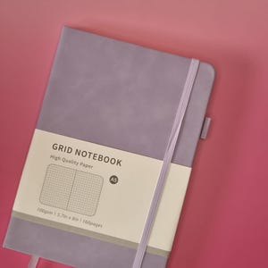 May include: A lavender A5 grid notebook with a matching elastic closure band and a paper belly band. The belly band features the text GRID NOTEBOOK, High Quality Paper, A5, 100gsm, 14.5cm x 20.3cm, and 160pages, along with a grid pattern graphic.