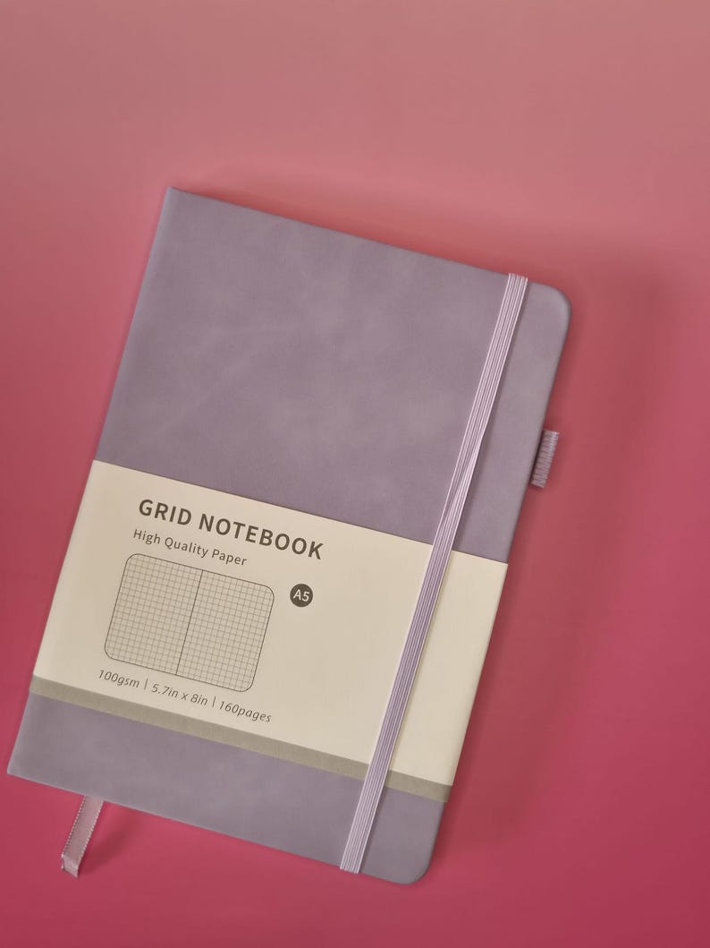 May include: A lavender A5 grid notebook with a matching elastic closure band and a paper belly band. The belly band features the text GRID NOTEBOOK, High Quality Paper, A5, 100gsm, 14.5cm x 20.3cm, and 160pages, along with a grid pattern graphic.