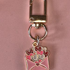 Puede incluir: Un llavero dorado con un mosquet&oacute;n y un peque&ntilde;o dije. El dije es un dise&ntilde;o de sobre rosa con un contorno dorado, lleno de corazones rojos, rosas y grises.