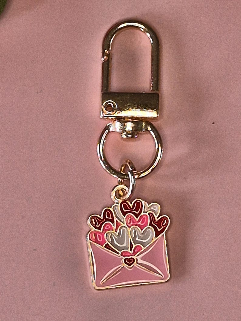 Puede incluir: Un llavero dorado con un mosquet&oacute;n y un peque&ntilde;o dije. El dije es un dise&ntilde;o de sobre rosa con un contorno dorado, lleno de corazones rojos, rosas y grises.