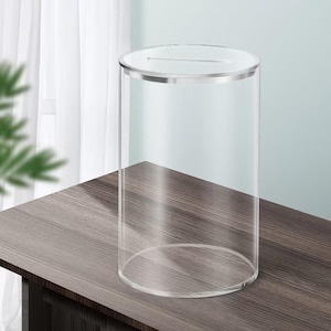 Op de afbeelding: Een transparante, cilindrische donatiebox met een gleuf aan de bovenkant. De box heeft een zilveren rand en staat op een donkerhouten oppervlak. De achtergrond omvat een wit gordijn en een lichtblauwe muur.