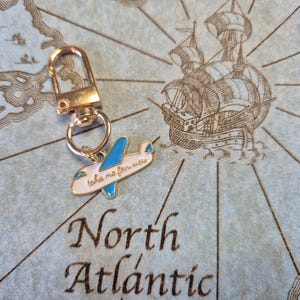 Puede incluir: Un llavero dorado con un peque&ntilde;o dije de avi&oacute;n. El avi&oacute;n es blanco con alas azules y el texto "take me far away". El llavero est&aacute; sobre un fondo de mapa con las palabras "North Atlantic".