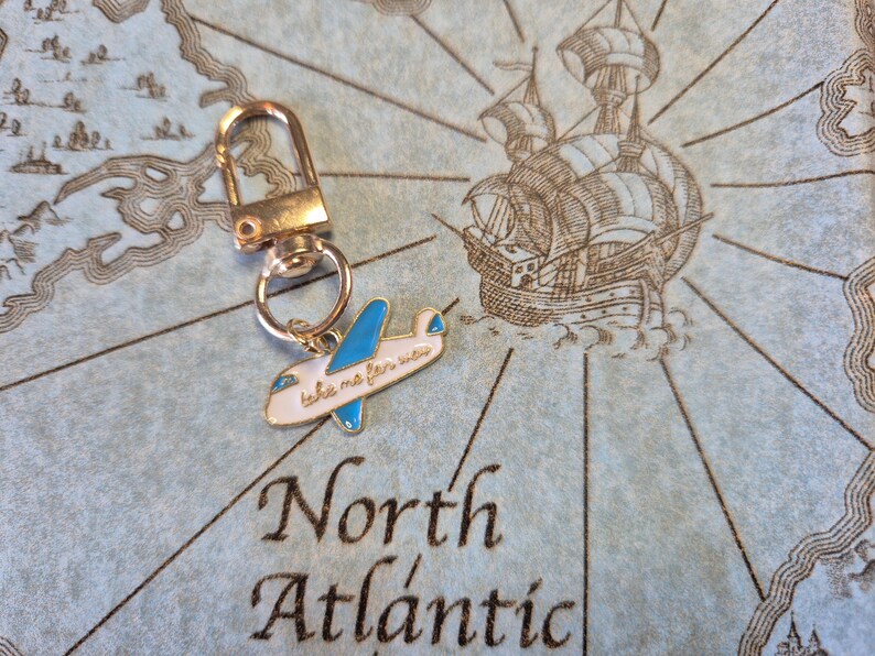 Puede incluir: Un llavero dorado con un peque&ntilde;o dije de avi&oacute;n. El avi&oacute;n es blanco con alas azules y el texto "take me far away". El llavero est&aacute; sobre un fondo de mapa con las palabras "North Atlantic".