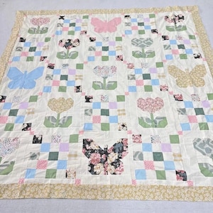 Könnte beinhalten: Eine handgefertigte Steppdecke mit einem sich wiederholenden Muster aus Blumen, Schmetterlingen und karierten Quadraten in Pastellfarben. Die Steppdecke hat einen cremefarbenen Hintergrund mit einem floralen Rand. Die Schmetterlinge sind rosa, blau und floral gemustert.