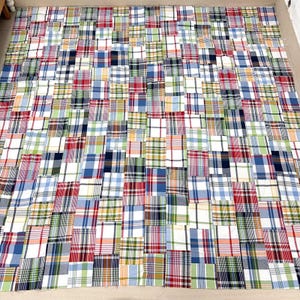 May include: A large patchwork quilt composed of numerous fabric squares in various plaid patterns. The quilt features a mix of colors, including red, blue, green, and white, creating a vibrant and textured appearance. The quilt is laid out flat.