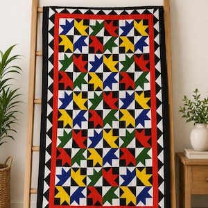 Könnte beinhalten: Ein hölzerner Leiterständer mit einem rechteckigen Quilt mit wiederkehrendem geometrischen Muster in Rot, Gelb, Blau, Grün, Schwarz und Weiß. Der Text oben lautet 01 HANG TO DISPLAY STAND. Perfect for showcasing your handmade quilt.