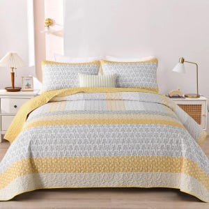 May include: A bed quilt set featuring a repeating pattern of gray and white floral motifs alternating with solid mustard yellow bands. The set includes two matching rectangular pillow shams and a central decorative pillow with vertical pleats.