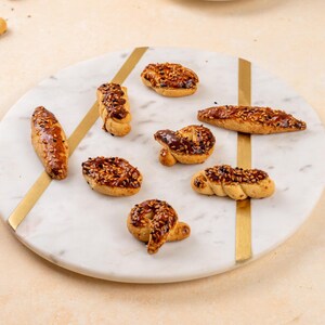 Op de afbeelding: Een assortiment gebakken deegwaren, waaronder verschillende vormen zoals krullen en ovalen, gepresenteerd op een marmeren serveerbord met gouden accenten. De goudbruine gebakjes zijn bedekt met sesamzaad. Een witte kop en schotel zijn zichtbaar op de achtergrond.