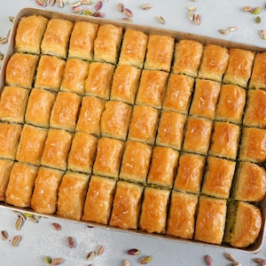 May include: A tray of golden-brown baklava pastries, arranged in neat rows. The dessert has a flaky, layered appearance, with visible layers of pastry and a glistening, syrup-soaked surface. Scattered pistachios add color and texture.