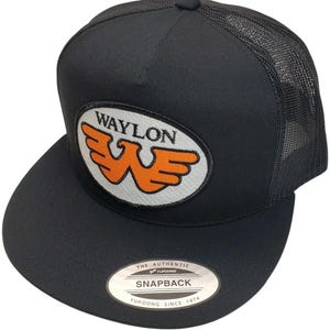 May include: Black trucker hat with a mesh back and a flat brim. The front features an oval patch with the name "WAYLON" above an orange "W" logo. The hat has a snapback closure and a "Yupoong" tag.