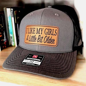 May include: Gray trucker hat with a black mesh back and a curved brim. The hat features a rectangular leather patch with the text "I LIKE MY GIRLS A Little Bit Older". The hat also has a Richardson patch.