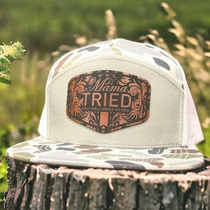 May include: A beige and white trucker hat with a camouflage pattern on the brim. The hat features a brown leather patch with the words "Mama Tried" and "Only Hell Mama Raised" in a decorative font.