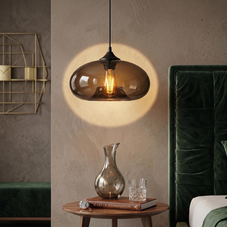 Op de afbeelding: Een moderne hanglamp met een rookglazen kap hangt aan een zwart snoer. De gloeilamp is zichtbaar in de kap. Een kleine tafel bevat een decoratieve vaas, een boek en een glas.