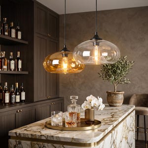 Op de afbeelding: Twee glazen hanglampen met amberkleurige en heldere kappen hangen boven een marmeren aanrecht. De lampen verlichten een bar met flessen drank, een dienblad met glazen en een vaas met witte bloemen. Het algemene thema is interieurdecoratie.
