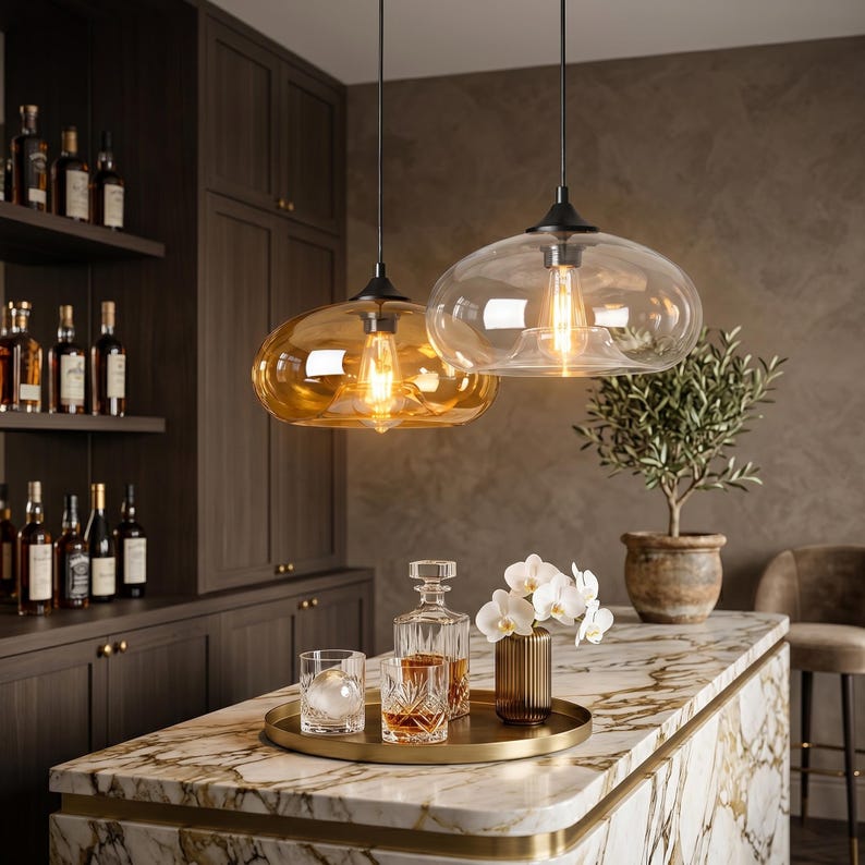 Op de afbeelding: Twee glazen hanglampen met amberkleurige en heldere kappen hangen boven een marmeren aanrecht. De lampen verlichten een bar met flessen drank, een dienblad met glazen en een vaas met witte bloemen. Het algemene thema is interieurdecoratie.