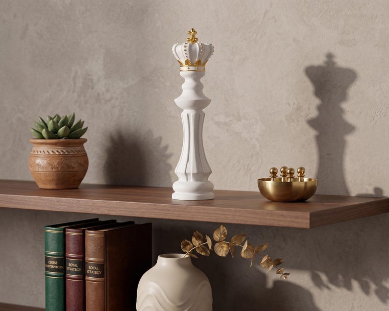 A white chess king figurine with a gold crown is on a wooden shelf next to a potted plant, books, and a small gold bowl.