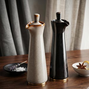 Two tall ceramic oil bottles, one speckled beige and one dark brown, stand on a wooden surface with a small dish of salt and lavender.