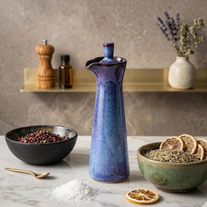 A tall, slender blue and purple glazed ceramic bottle with a fluted spout stands on a white marble surface, surrounded by bowls of spices and dried citrus.
