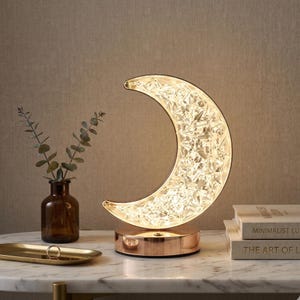 May include: A crescent moon-shaped table lamp with a copper base and a sparkling, textured surface. The lamp is illuminated, casting a warm glow. A small vase with greenery and books are nearby.