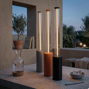 May include: Three modern outdoor lamps with cylindrical bases in gray, terracotta, and black. Each lamp has a clear glass tube with a vertical light. A glass carafe, a bowl of ice, and a pencil are on the stone table.
