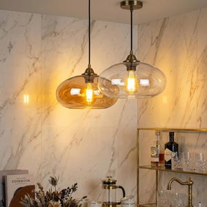 Op de afbeelding: Twee hanglampen met heldere en amberkleurige glazen kappen hangen aan het plafond. De lampen hebben een vintage messing afwerking en zichtbare Edison-stijl lampen. De achtergrond heeft een marmerpatroon muur en een messing plank.