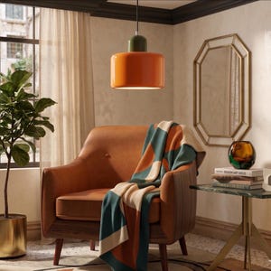 Peut inclure: Un luminaire suspendu orange et vert éclaire un fauteuil en cuir marron. Une couverture à motifs géométriques, dans les tons de teal, orange et crème, est drapée sur le fauteuil. Un miroir doré et une table d'appoint avec des livres et un vase complètent le décor.
