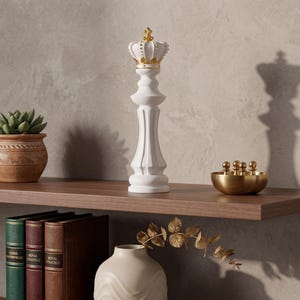 May include: A white chess piece in the shape of a king, topped with a gold crown, stands on a wooden shelf. A small succulent in a terracotta pot and a gold bowl with small spheres are also on the shelf.