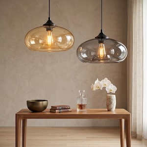 Op de afbeelding: Twee hanglampen met ovale, amberkleurige en grijze glazen kappen hangen boven een houten tafel. De tafel is versierd met een messing kom, boeken, een karaf en een marmeren vaas met witte bloemen. De lampen hebben zwarte armaturen.