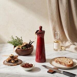 A red ceramic oil bottle stands on a marble surface next to a wooden bowl with herbs and vanilla beans, a small dish of star anise, and a plate with a slice of bread and a knife.