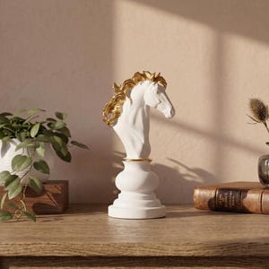 May include: A white horse head bust with gold accents on the mane and a gold band around the neck. The bust sits on a white pedestal base. The sculpture is displayed on a wooden surface with a plant and books in the background.