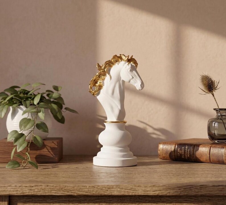 May include: A white horse head bust with gold accents on the mane and a gold band around the neck. The bust sits on a white pedestal base. The sculpture is displayed on a wooden surface with a plant and books in the background.