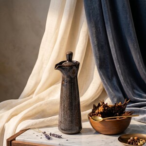 May include: A dark brown ceramic oil dispenser with a lid and spout. A wooden bowl filled with spices and a gold spoon are placed nearby. The background features draped fabric in cream and navy blue.