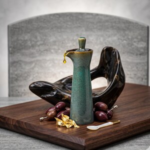 May include: A teal ceramic oil dispenser with a small spout, dripping oil. The dispenser is on a wooden cutting board with olives, a decorative spoon, and a gold leaf. A dark wooden sculpture is behind the dispenser.