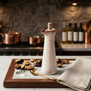 May include: A cream ceramic oil cruet with a brown rim and stopper. The cruet is on a wooden tray with a marble insert, with a gold spoon, mushrooms, and a linen napkin.
