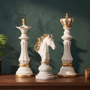 Three white chess pieces, a queen, knight, and king, with gold accents, stand on a dark wooden surface next to a green plant and a mirror.