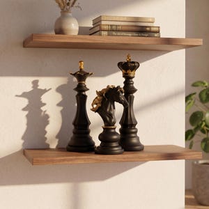 May include: Two wooden shelves display decorative items. The lower shelf holds three black chess pieces with gold accents. The upper shelf features a vase with dried flowers and a stack of books. The shelves are mounted on a white wall.