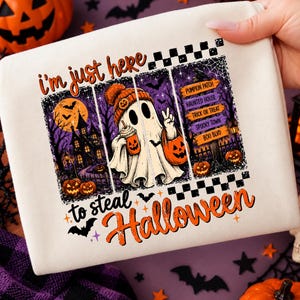 May include: A white fabric item featuring a graphic print with the text 'i'm just here to steal Halloween'. The design includes a ghost wearing an orange beanie, holding a drink and a pumpkin bucket, alongside a haunted house and signposts.