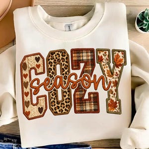 May include: A cream-colored crewneck sweatshirt featuring the word COZY in large block letters with varied patterns including hearts, leopard print, plaid, and autumn leaves, with the word season in cursive script layered over the center.