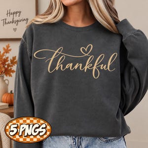 May include: A charcoal gray crewneck sweatshirt featuring the word thankful in a light tan cursive script with a small heart icon above the letter k. The garment has long sleeves and a ribbed neckline, providing a simple and clean appearance.