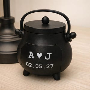 May include: A matte black cauldron-shaped container with a lid and a handle. The front features white text displaying the letters A and J separated by a heart symbol, with the date 02.05.27 printed in white below the letters on the rounded body.