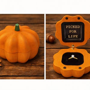 May include: A pumpkin-shaped ring box with a matte orange exterior and a green stem. The box opens to reveal a black interior lining and a gold ring with a clear stone. The inside lid features the gold text PICKED FOR LIFE in a square frame.