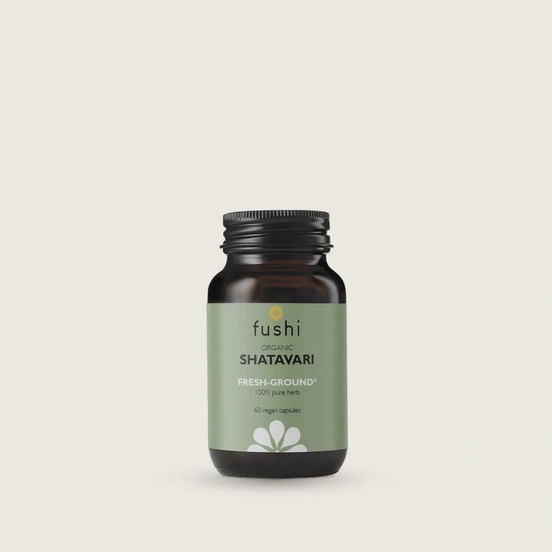 May include: A dark brown glass bottle with a black screw-top lid containing 60 vegan capsules. The label is sage green with the text Fushi, Organic Shatavari, Fresh-Ground, 100% pure herb, and 60 vegan capsules, featuring a white flower icon.