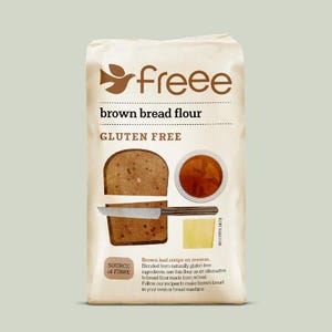 May include: A cream-coloured paper bag of Freee brown bread flour. The front features the brand logo, the text "GLUTEN FREE," an illustration of sliced brown bread with a knife, a small bowl of liquid, a square of cheese, and descriptive text.