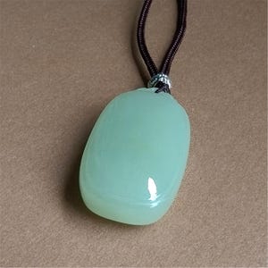 May include: A light green, oval-shaped pendant necklace. The pendant is smooth and glossy, with a subtle, translucent quality. It hangs from a dark brown cord. The necklace is a simple, elegant piece of jewelry.