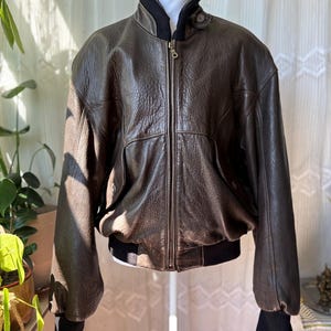 Puede incluir: Chaqueta bomber de cuero marrón con cuello y puños negros. La chaqueta tiene cierre de cremallera y dos bolsillos delanteros. El cuero tiene un aspecto texturizado. La chaqueta se exhibe en un maniquí blanco.
