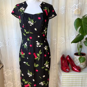 Puede incluir: Vestido negro con estampado de cerezas y flores. El vestido presenta un escote cuadrado, mangas cortas y una silueta entallada. Zapatos de tacón rojos peep-toe se colocan junto al vestido sobre una mesa a rayas.
